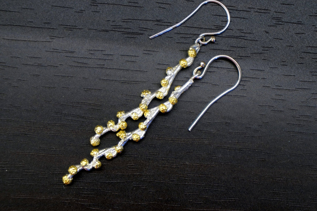 A pair of silver earrings with a unique design, featuring a long, thin branch-like structure with small, round, gold-colored beads protruding from it.