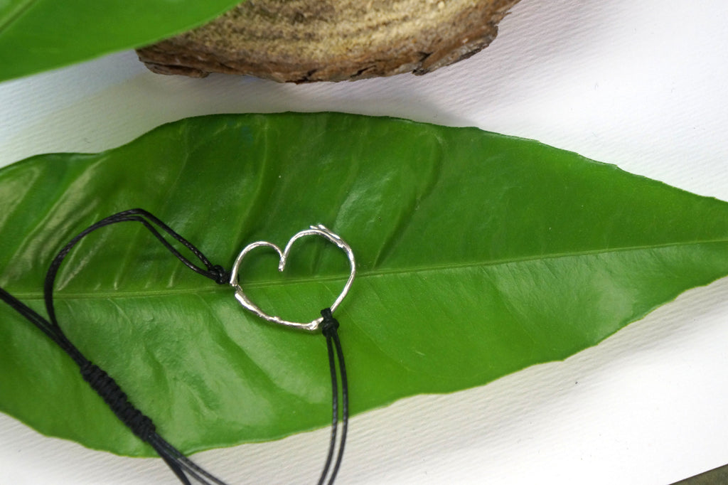 A silver heart-shaped charm is attached to a black string, resting on a large green leaf.