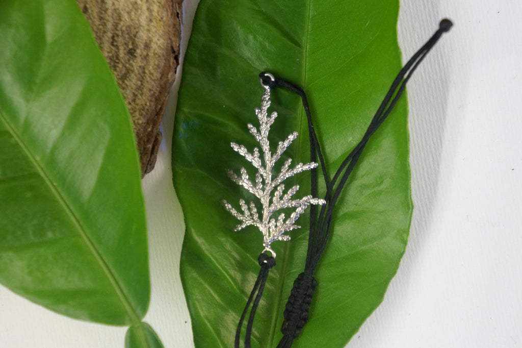A silver leaf-shaped pendant with a black cord is displayed on a green leaf against a white background.