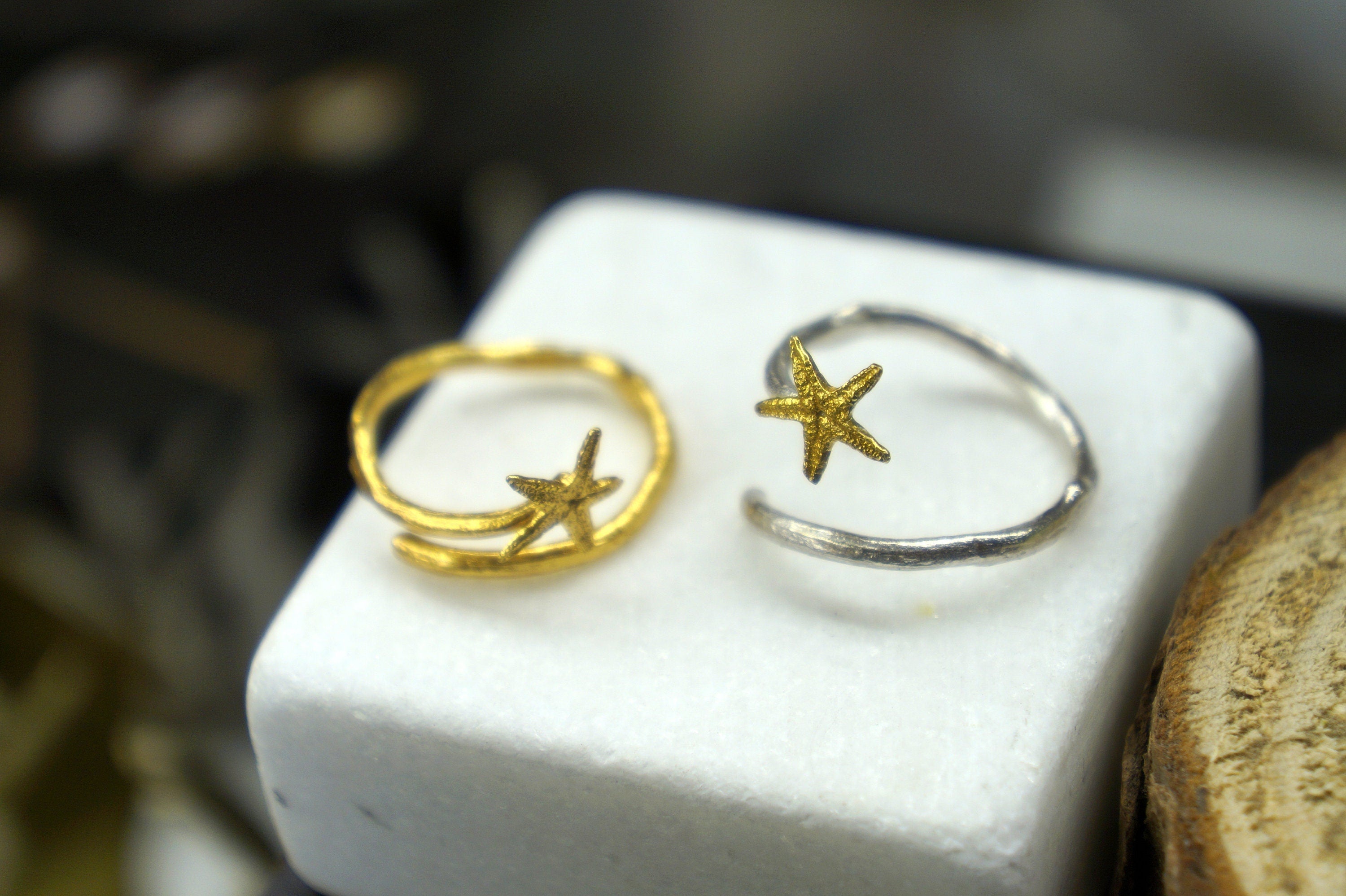 Two gold star-shaped earrings are displayed on a white pedestal, one on each side of a silver star-shaped earring.