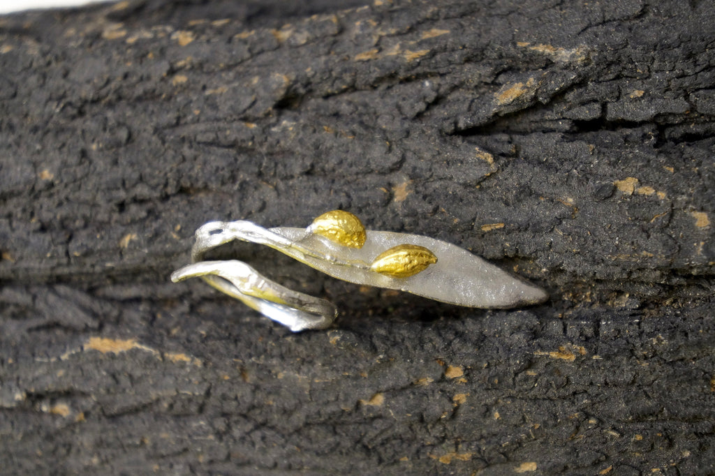 A close-up image of a leaf-shaped piece of jewelry, possibly a ring, with two gold-colored leaves attached to it, resting on a textured, dark surface.