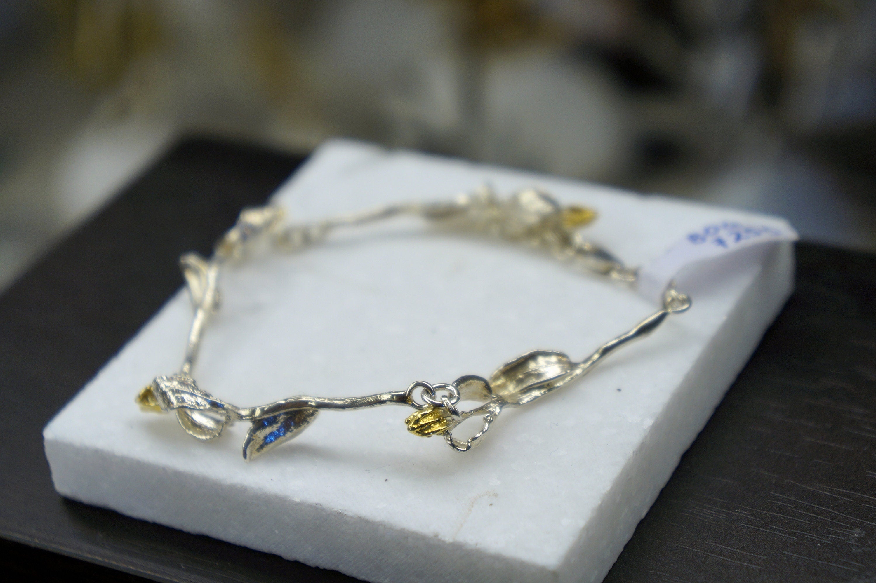 A silver bracelet with leaf-shaped charms and a tag is displayed on a white surface.