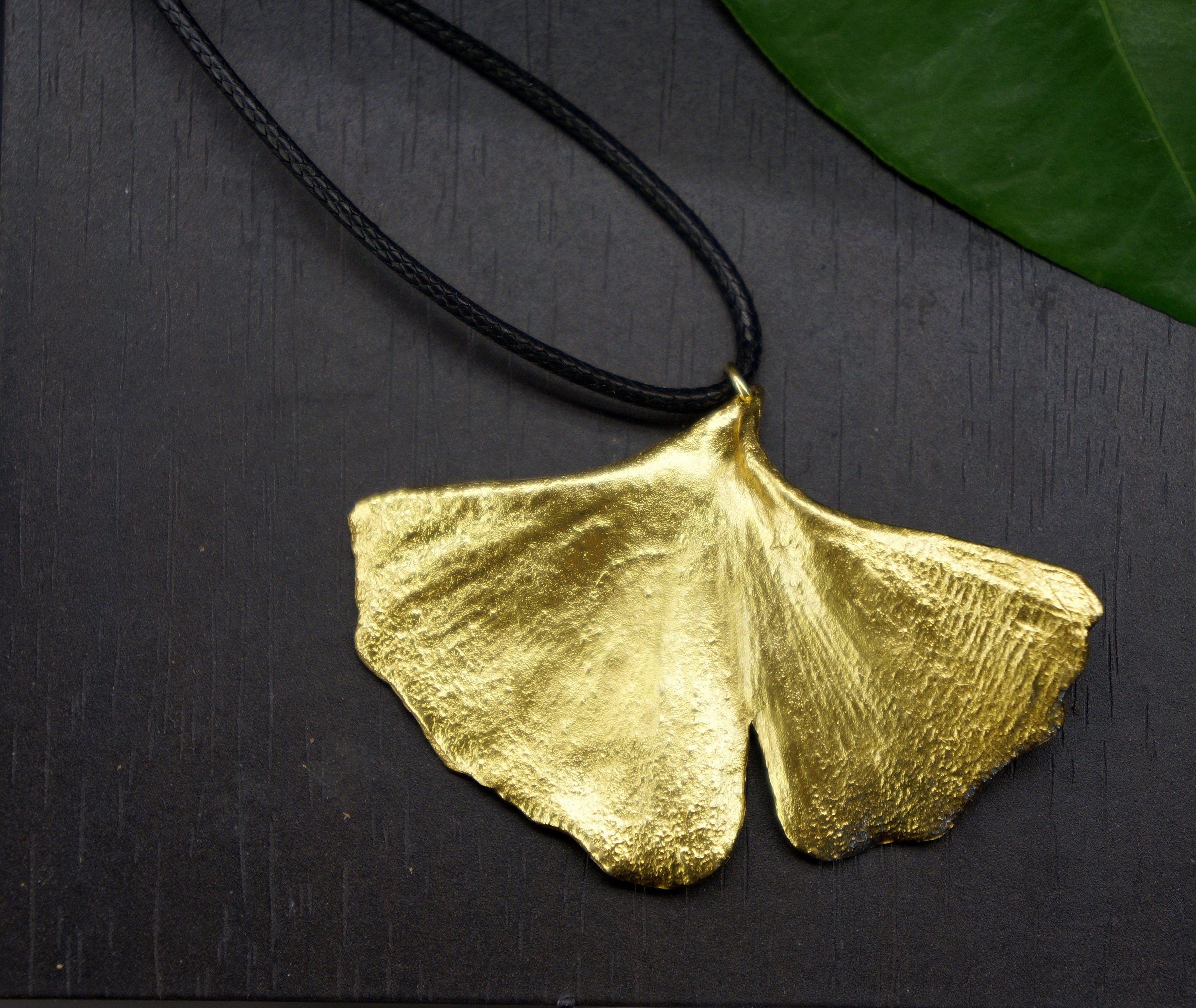 A gold leaf pendant with a black cord is displayed on a dark surface, with a green leaf visible in the background.