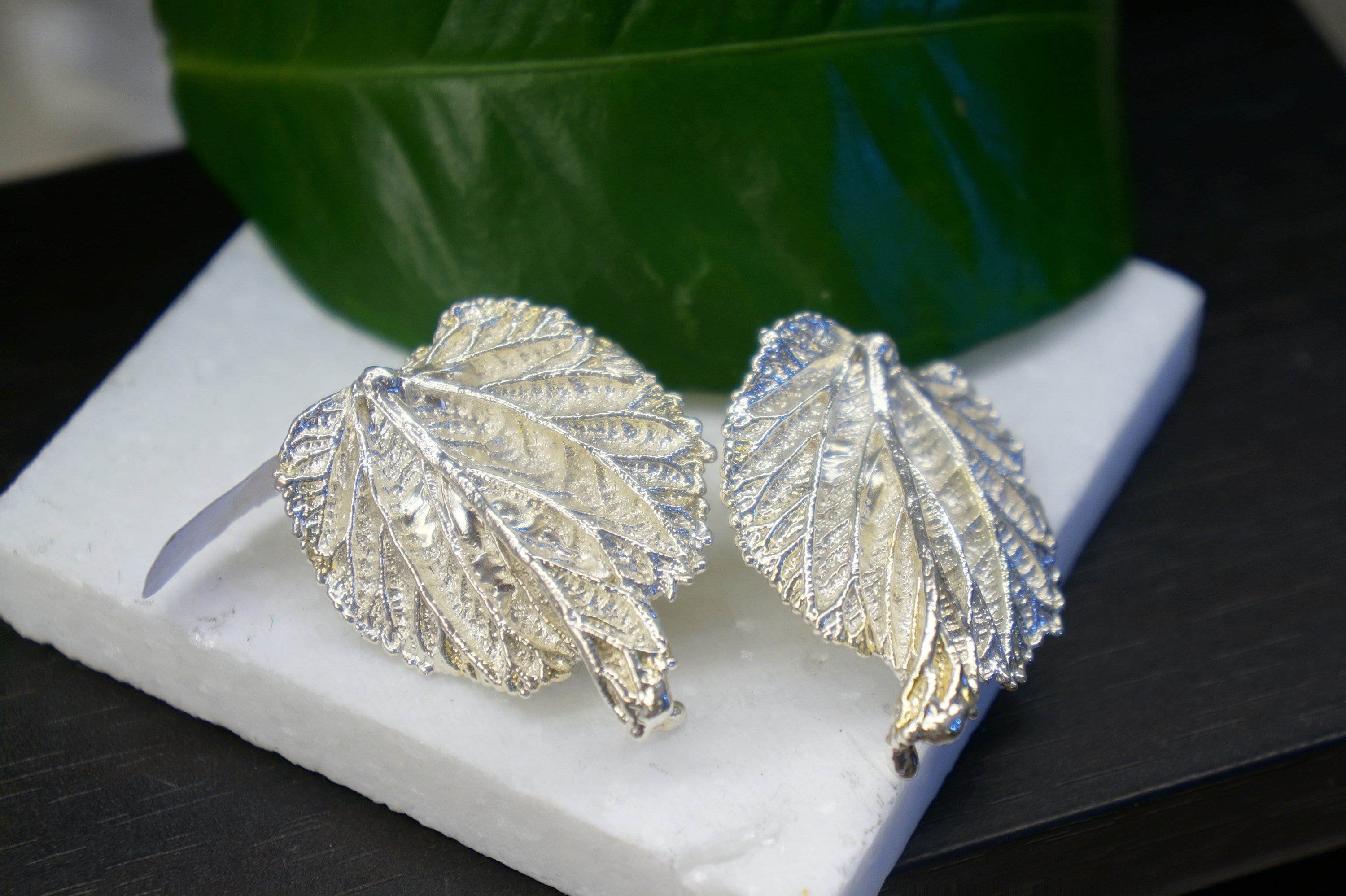 two silver leaf-shaped earrings placed on a white surface, with a green leaf in the background.