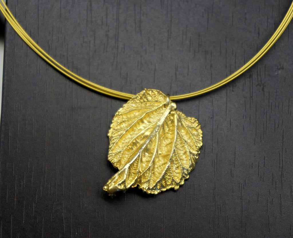 A gold leaf pendant with a thin gold chain is displayed against a dark background.