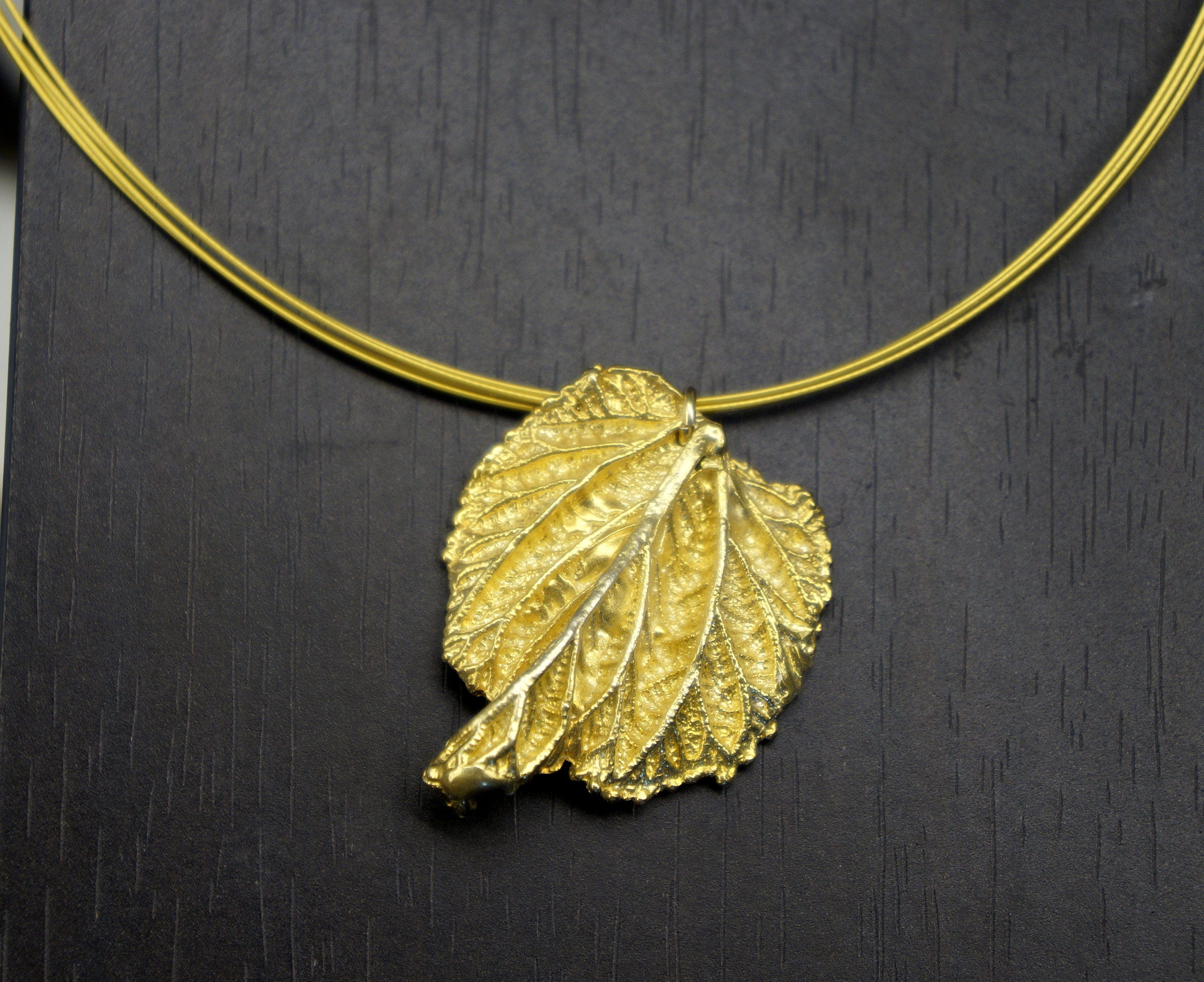 A gold leaf pendant with a thin gold chain is displayed against a dark background.