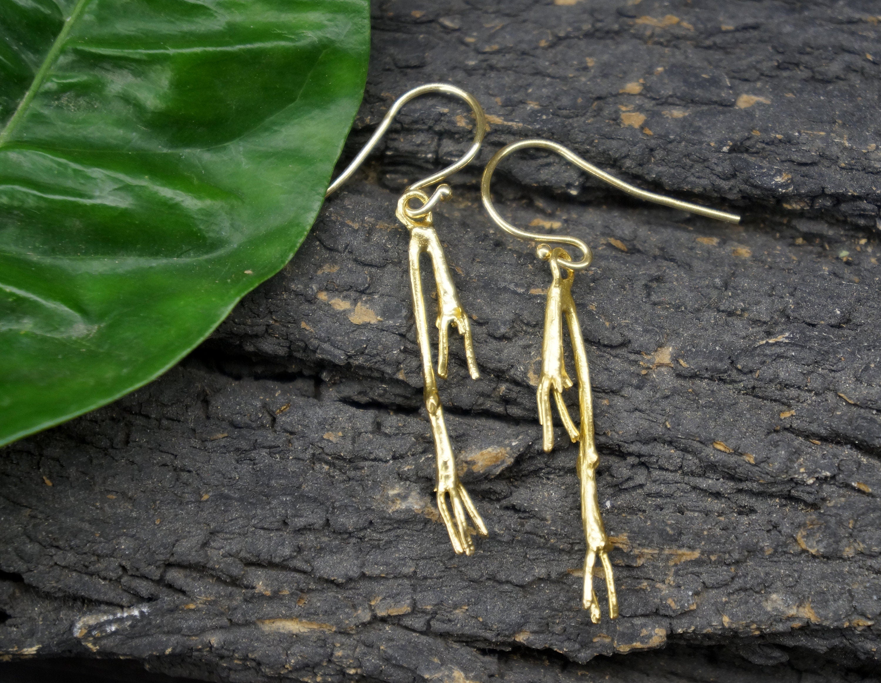 A pair of gold earrings with a leaf-like design rests on a dark surface, with the leaf's green color contrasting with the dark background.