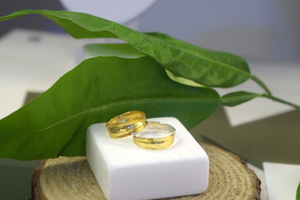 Two gold wedding rings are placed on a white pedestal, surrounded by a large green leaf and a wooden log, creating a natural and elegant setting.