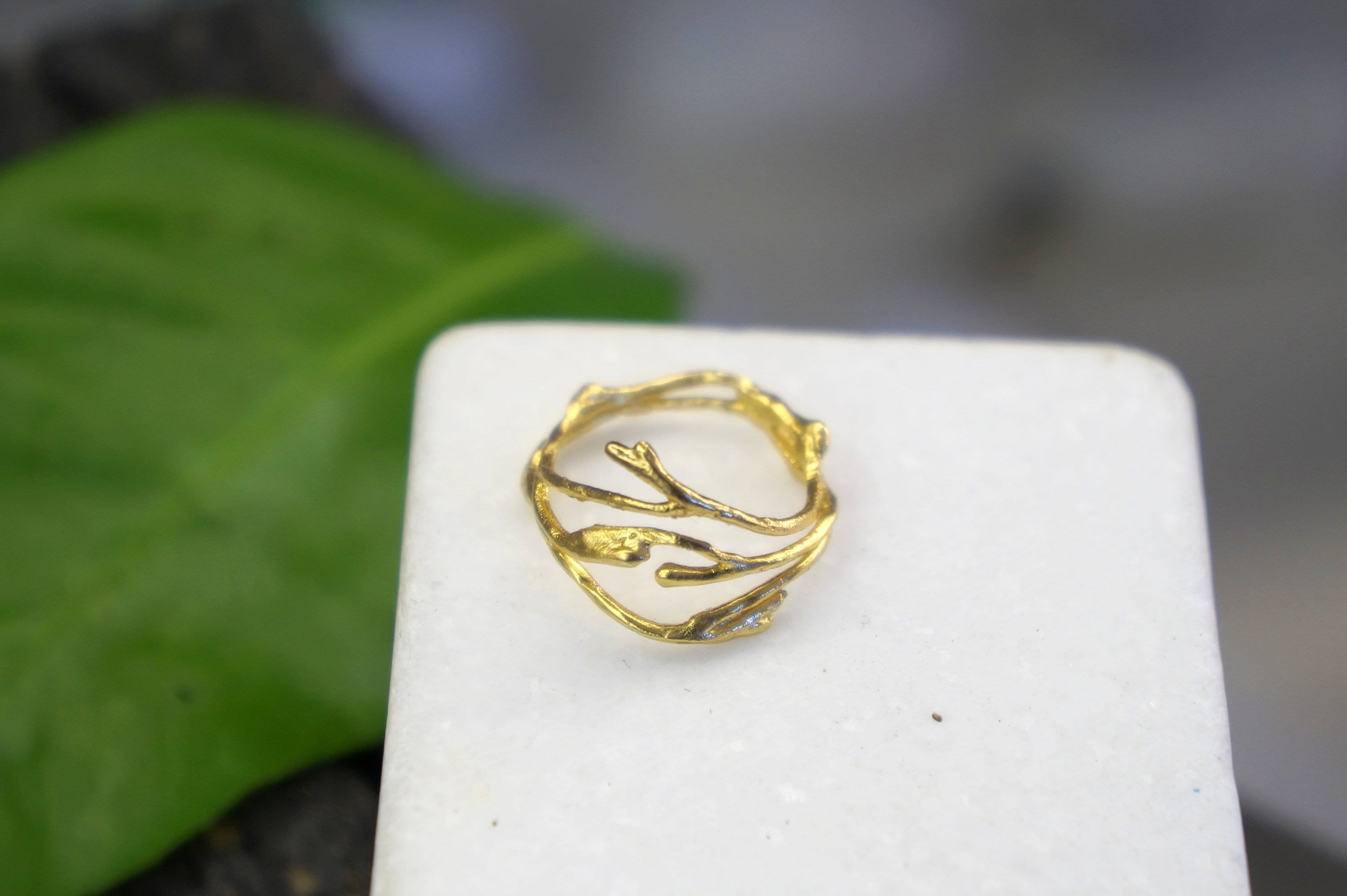A gold ring with a leaf-like design sits on a white surface, with a blurred green leaf in the background.