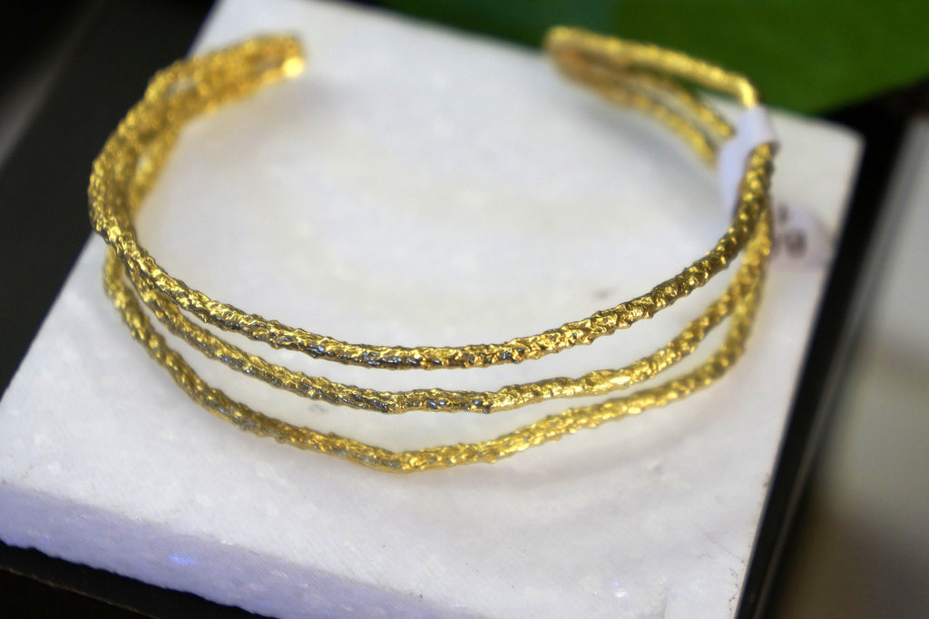 A close-up image of a gold bracelet with a textured, three-strand design, resting on a white surface.