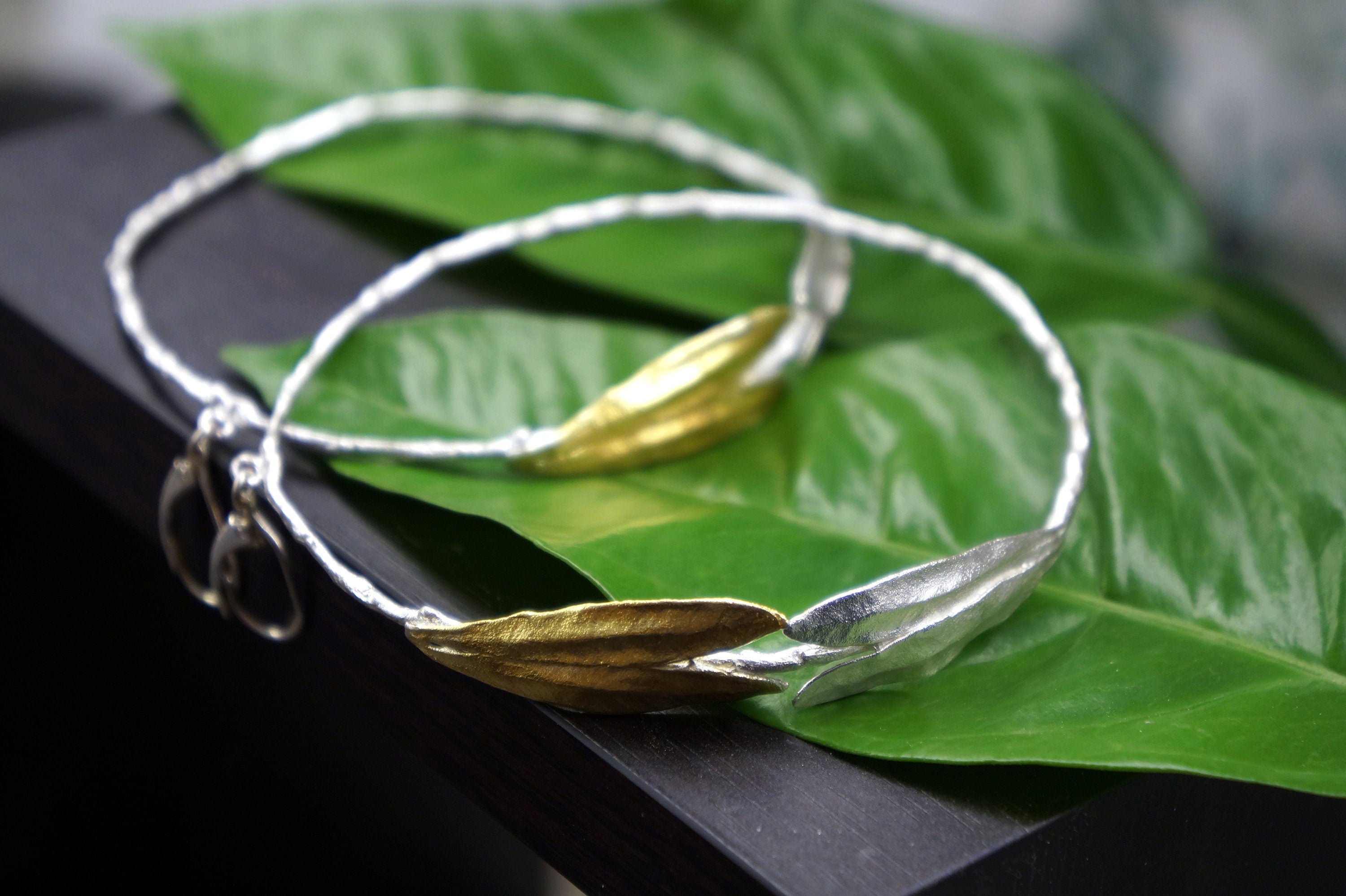 A pair of silver and gold leaf-shaped bracelets resting on a green leaf.