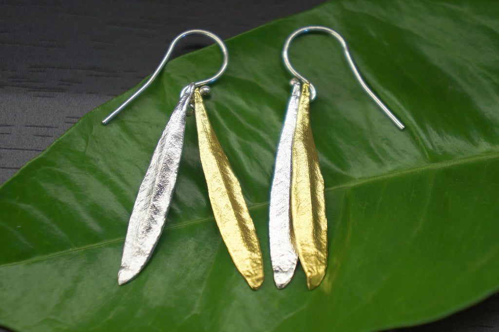 A pair of earrings with gold and silver leaf-like designs resting on a green leaf.