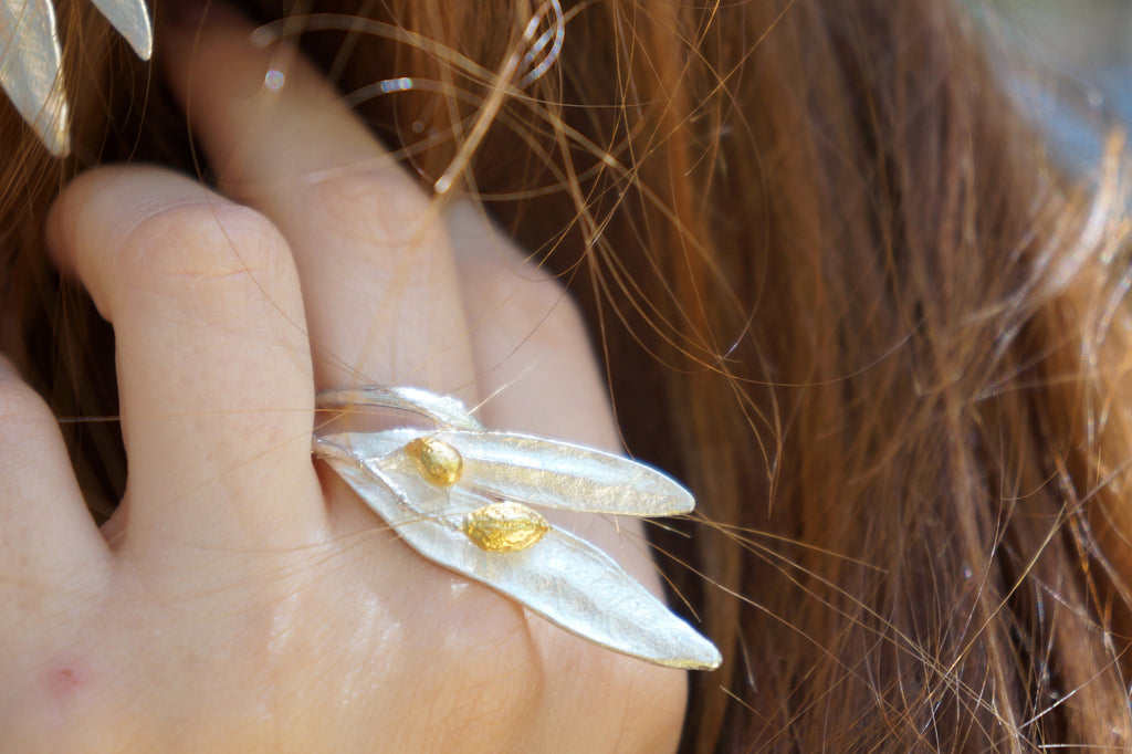 A close-up of a person's hand wearing a silver leaf-shaped ring with two gold leaves.