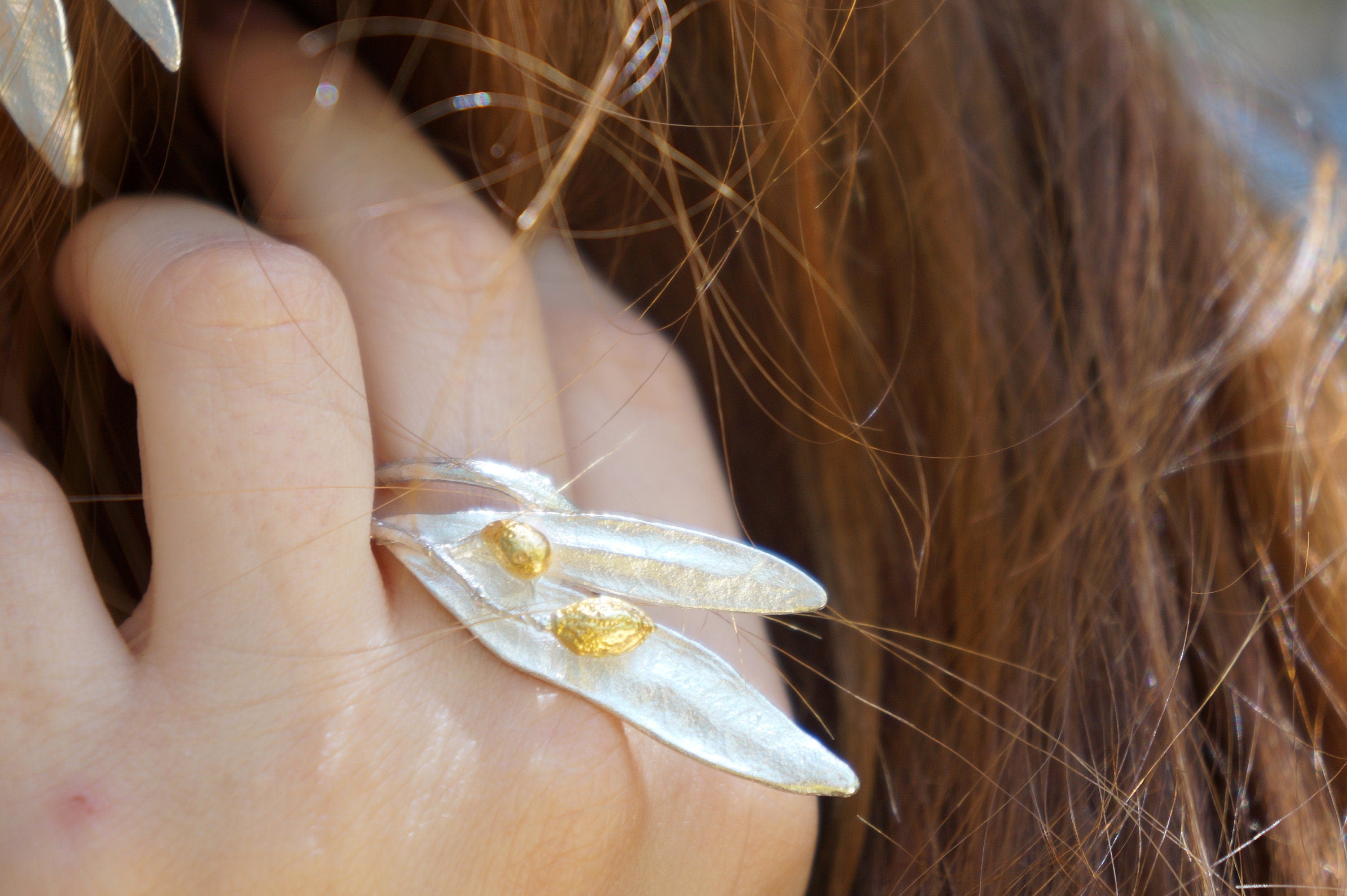 A close-up of a person's hand wearing a silver leaf-shaped ring with two gold leaves.