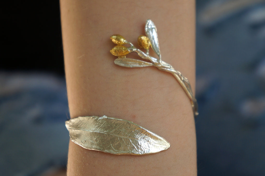 a person's arm wearing a silver leaf-shaped bracelet and a small gold leaf-shaped charm on the wrist.