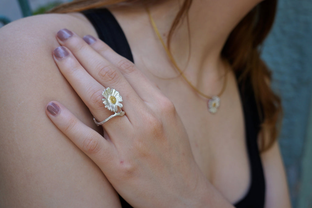 A close-up of a person's hand wearing a ring with a flower design, and a necklace around their neck.
