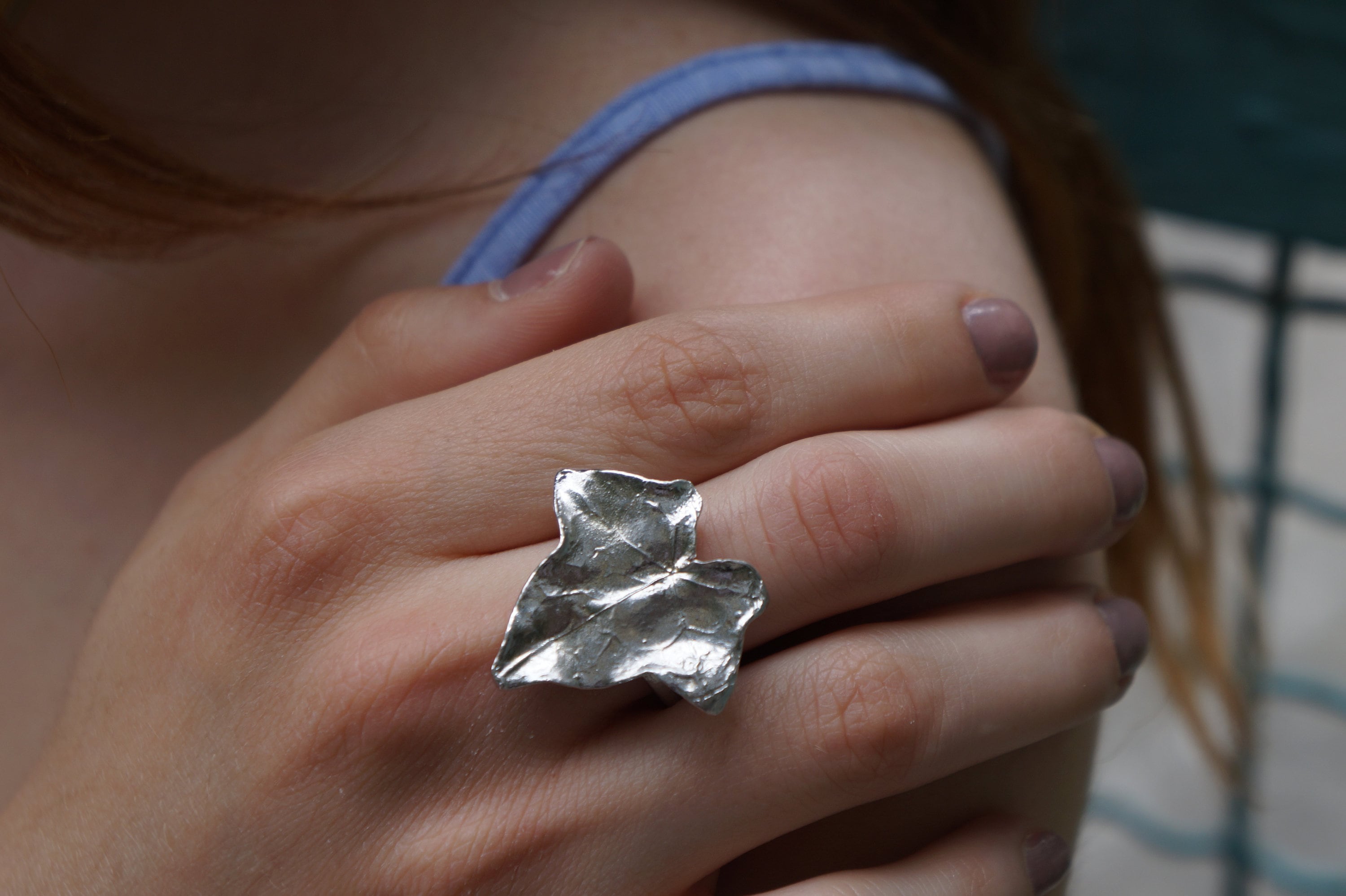 A close-up of a person's hand wearing a silver leaf-shaped ring, with a blue top visible in the background.