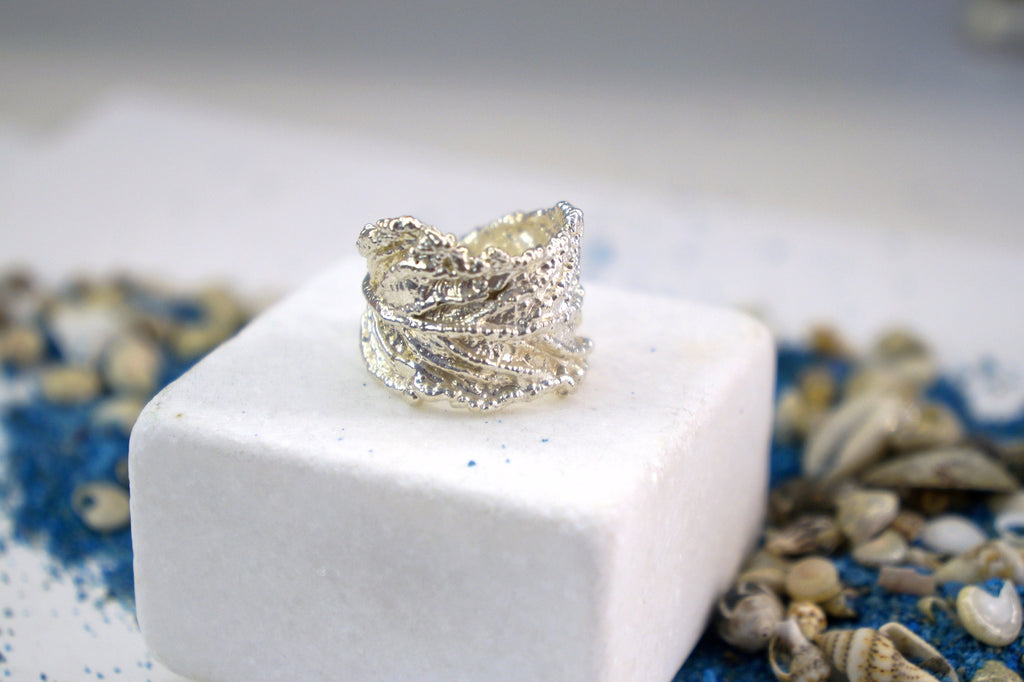 A silver ring with a textured, leaf-like design sits atop a white square base, surrounded by scattered seashells and blue sand.