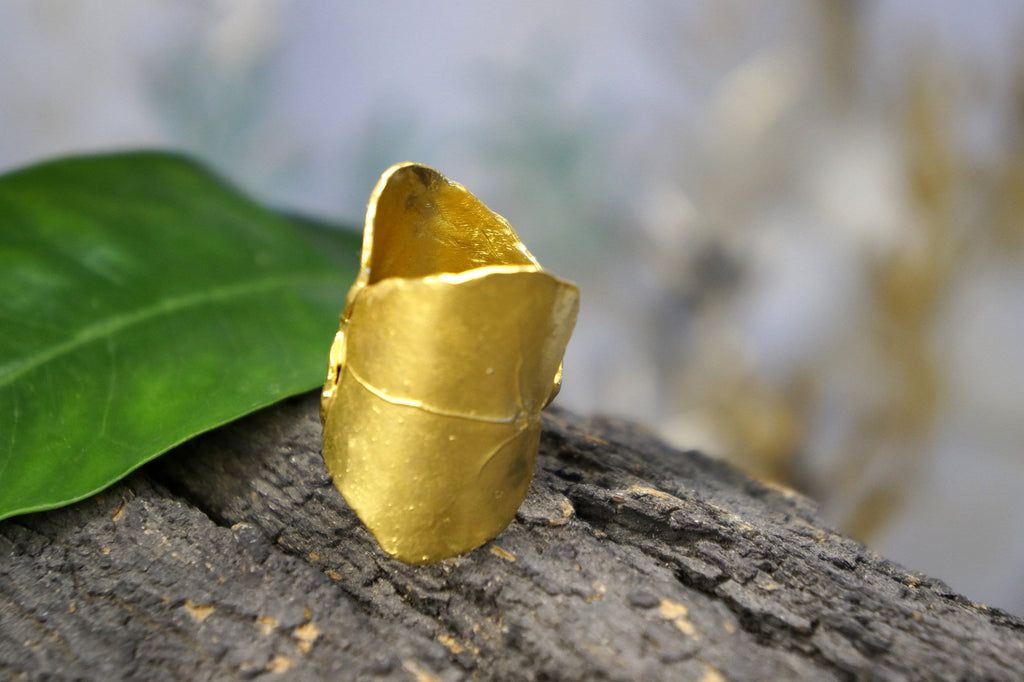 A gold ring with a unique shape sits on a wooden surface, with a blurred green leaf in the background.
