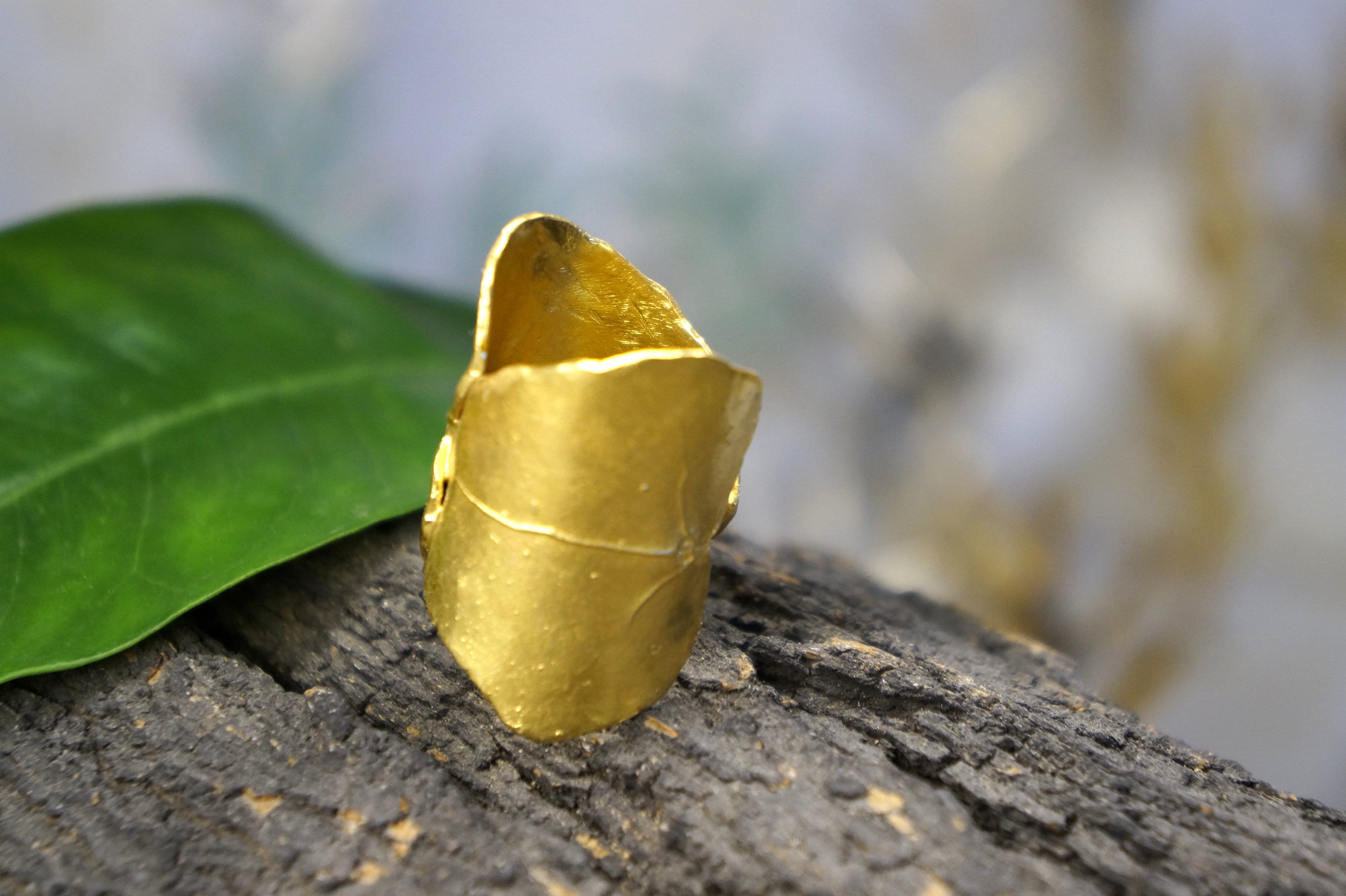 A gold ring with a unique shape sits on a wooden surface, with a blurred green leaf in the background.