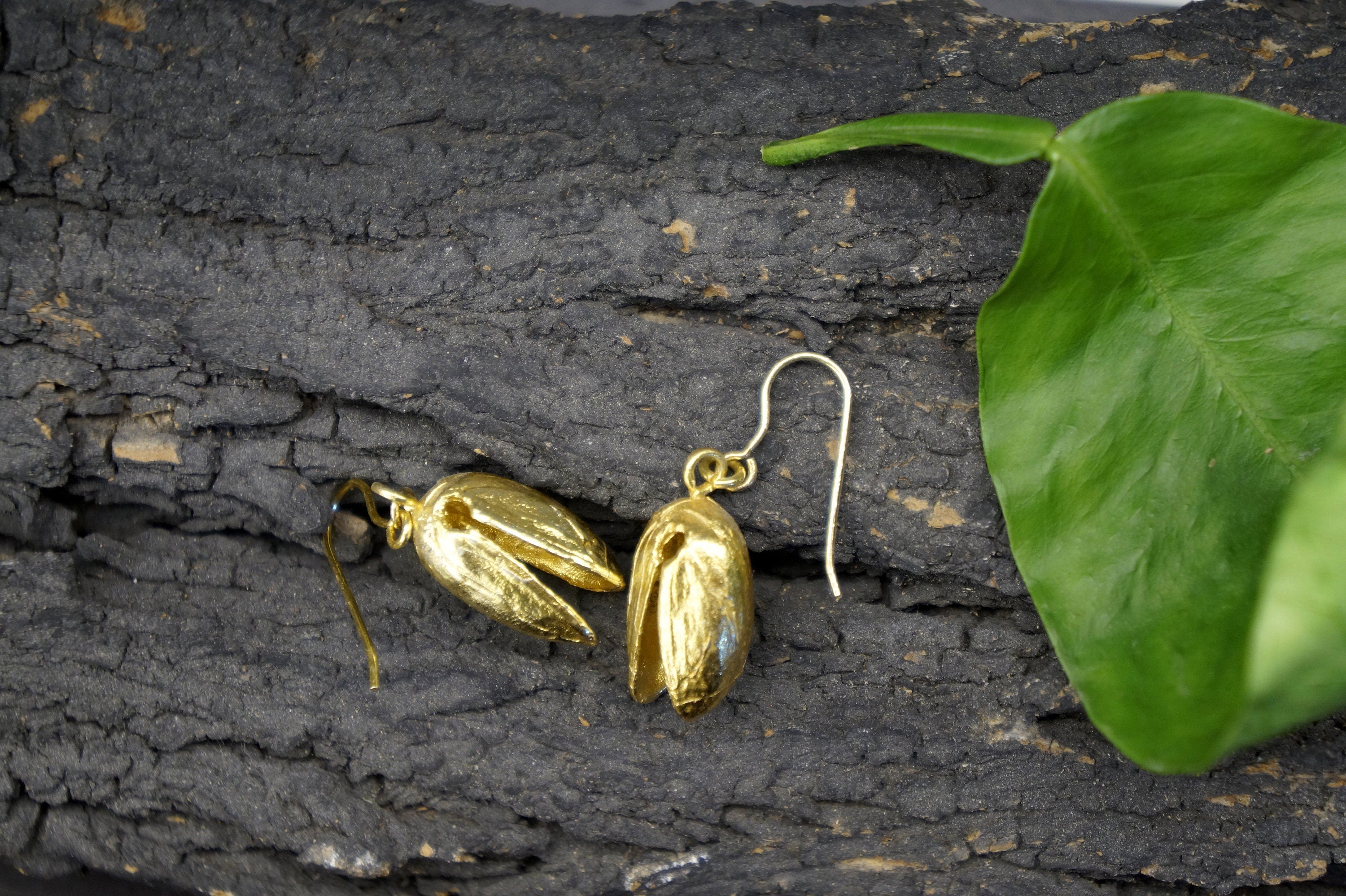 A pair of gold earrings with a leaf-like design rests on a textured surface, accompanied by a green leaf.