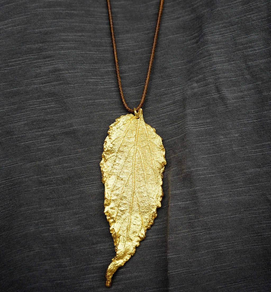 A gold leaf pendant with a brown cord is displayed against a dark gray background.