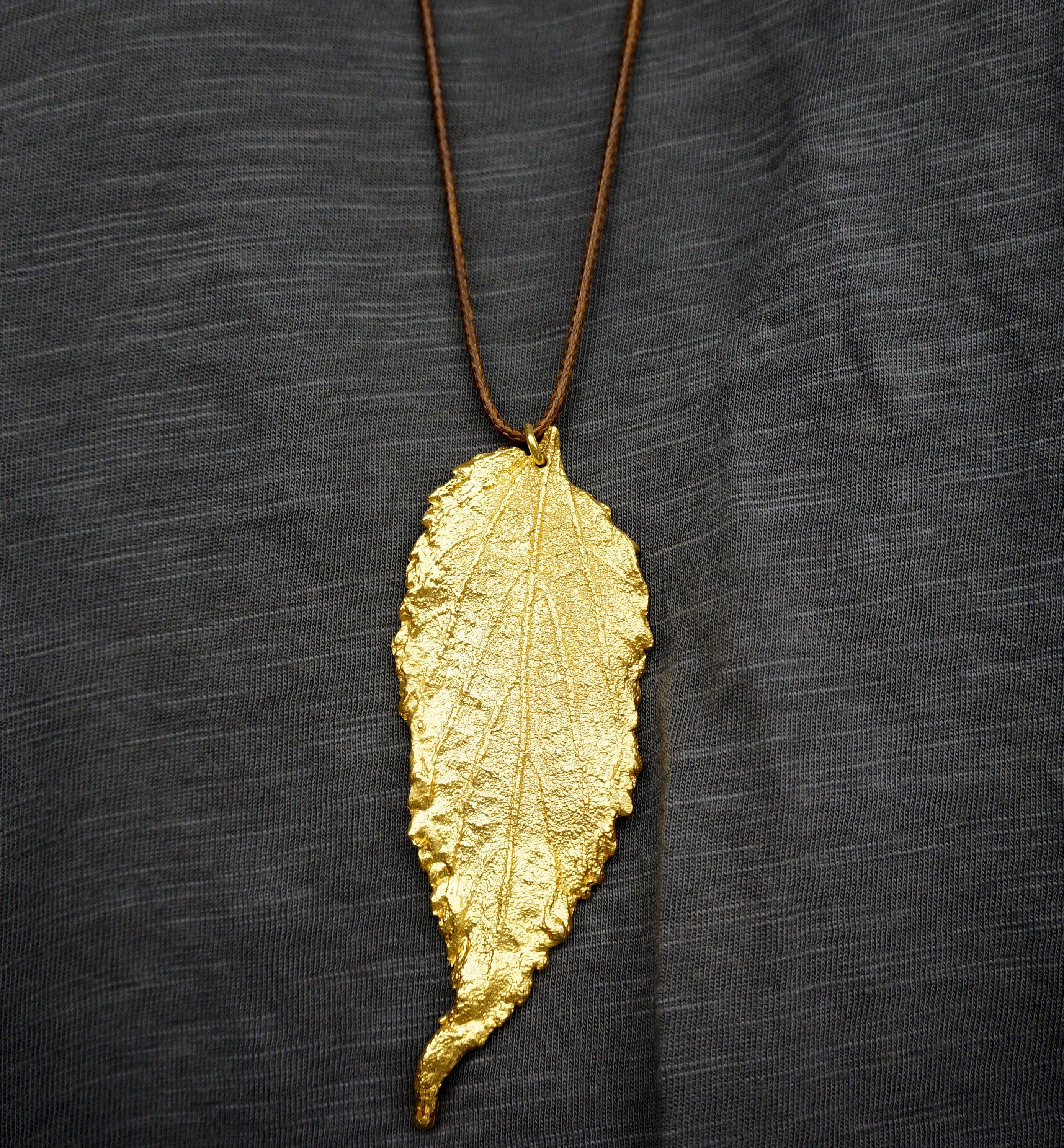 A gold leaf pendant with a brown cord is displayed against a dark gray background.