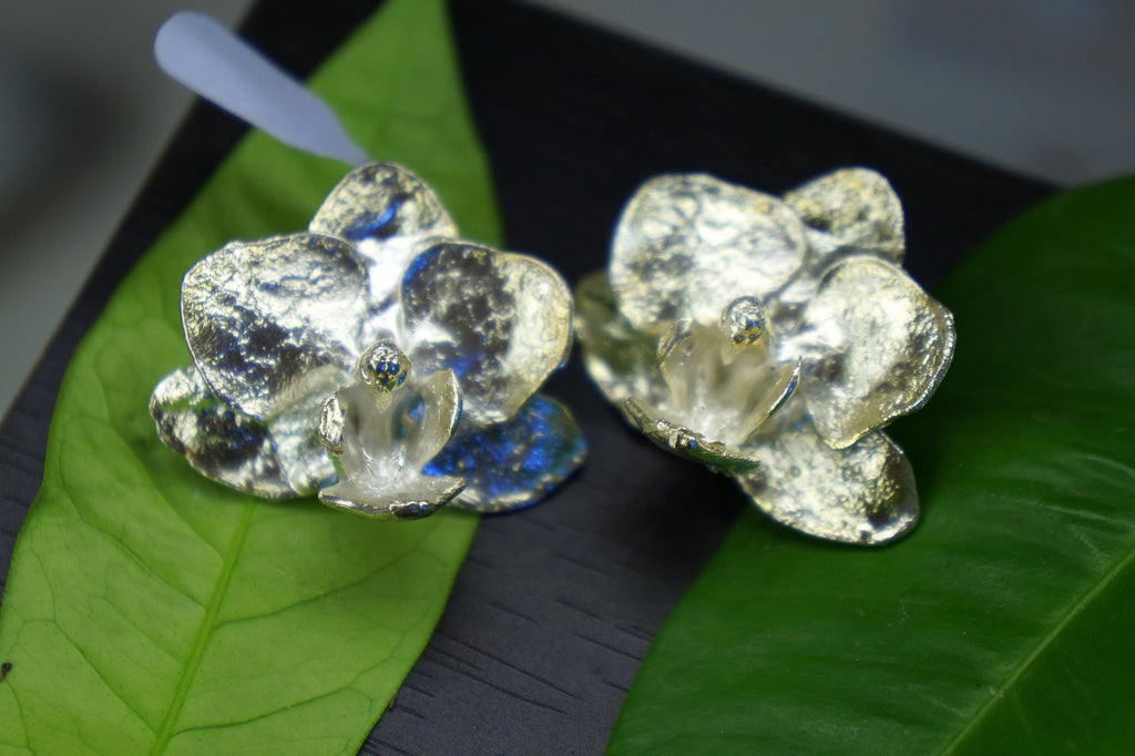 Two silver orchid-shaped earrings are displayed on a green leaf, with a white object in the background.