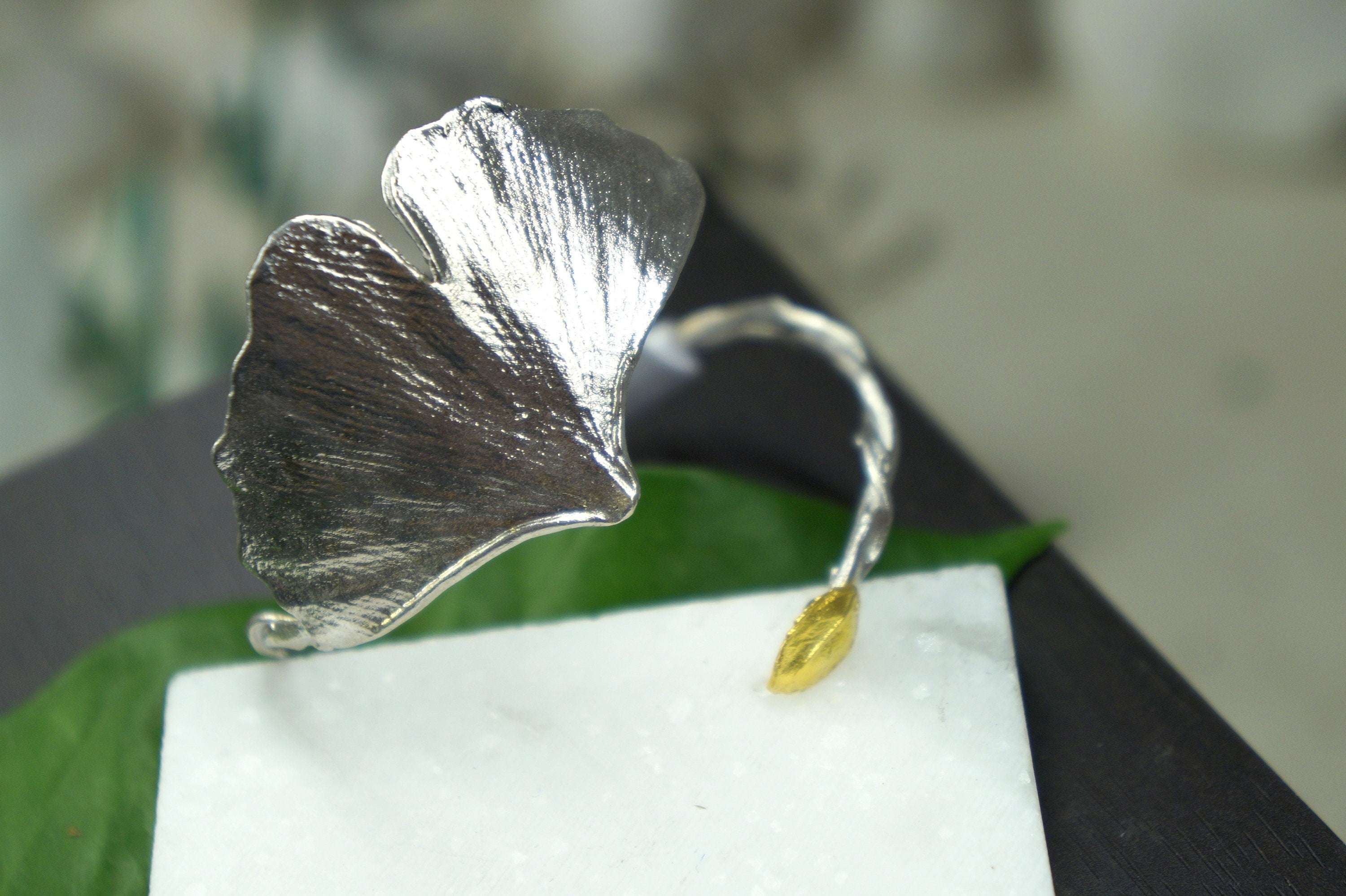 A close-up view of a silver leaf-shaped ring with a gold accent, resting on a white surface.