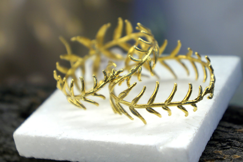 two gold leaf-like bracelets with intricate designs, resting on a white foam base.