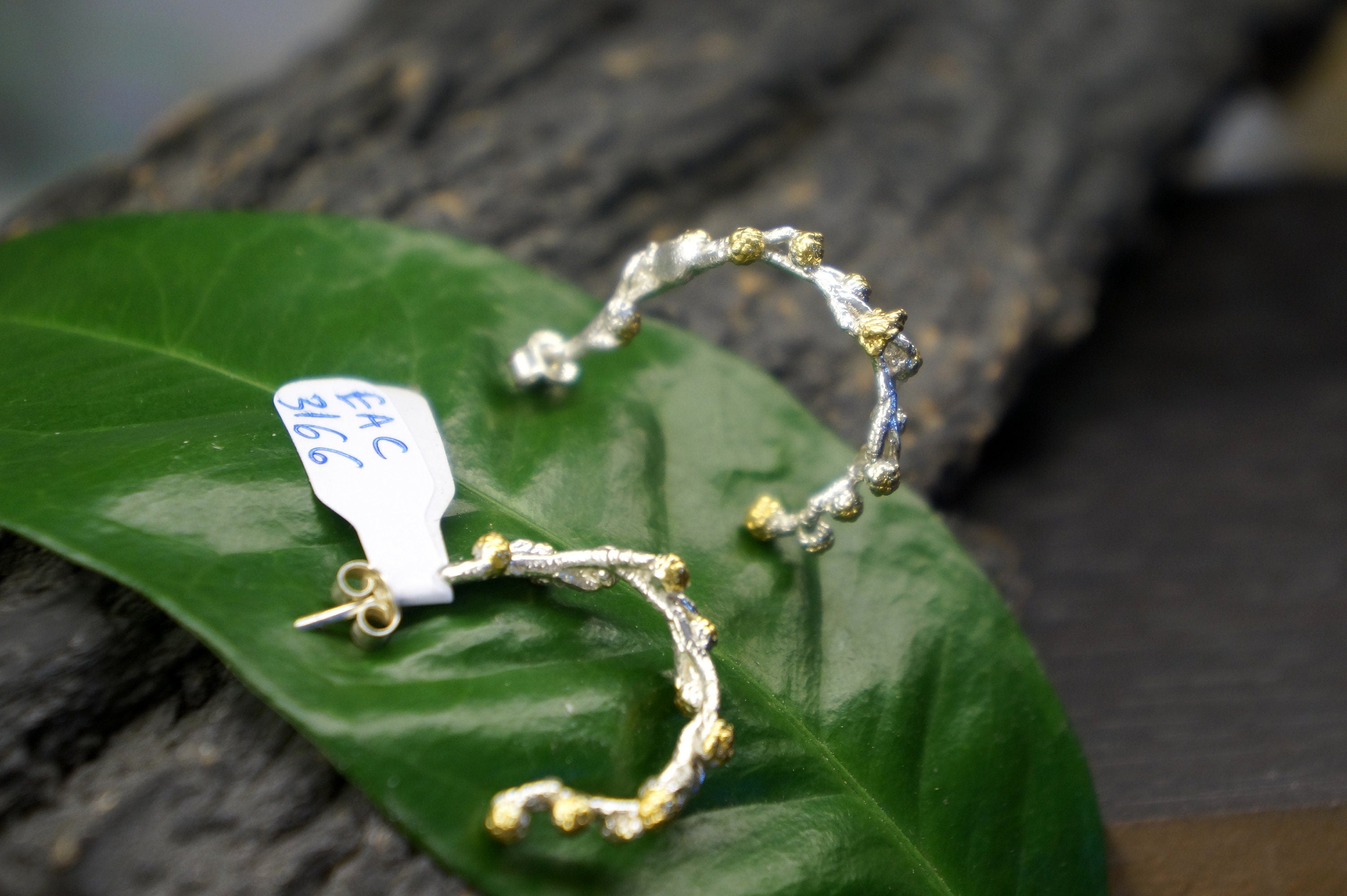 A pair of silver earrings with a unique, organic design rests on a green leaf.