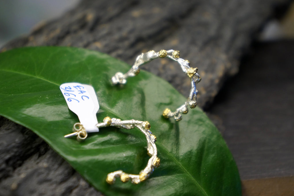 A pair of silver earrings with a unique, organic design rests on a green leaf.