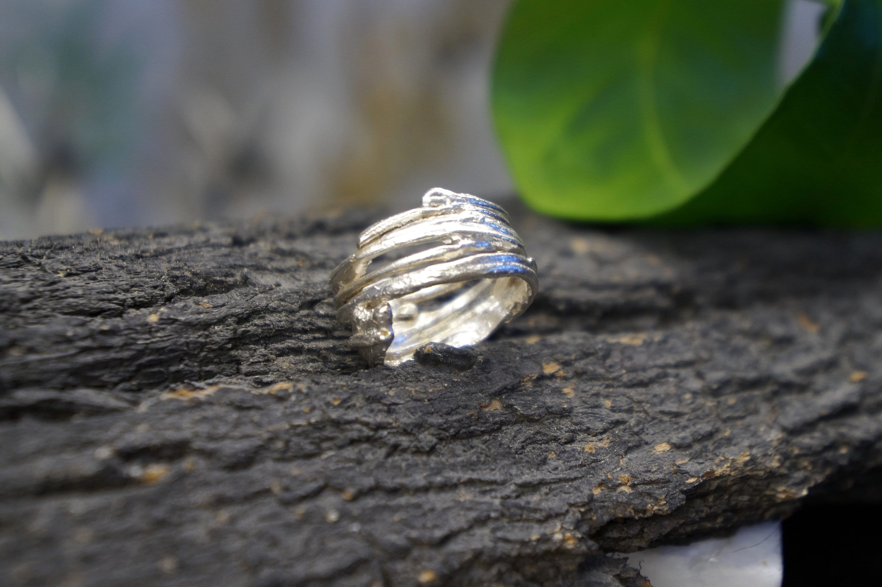 A silver ring with a unique, twisted design rests on a rough, textured surface, with a blurred green leaf in the background.