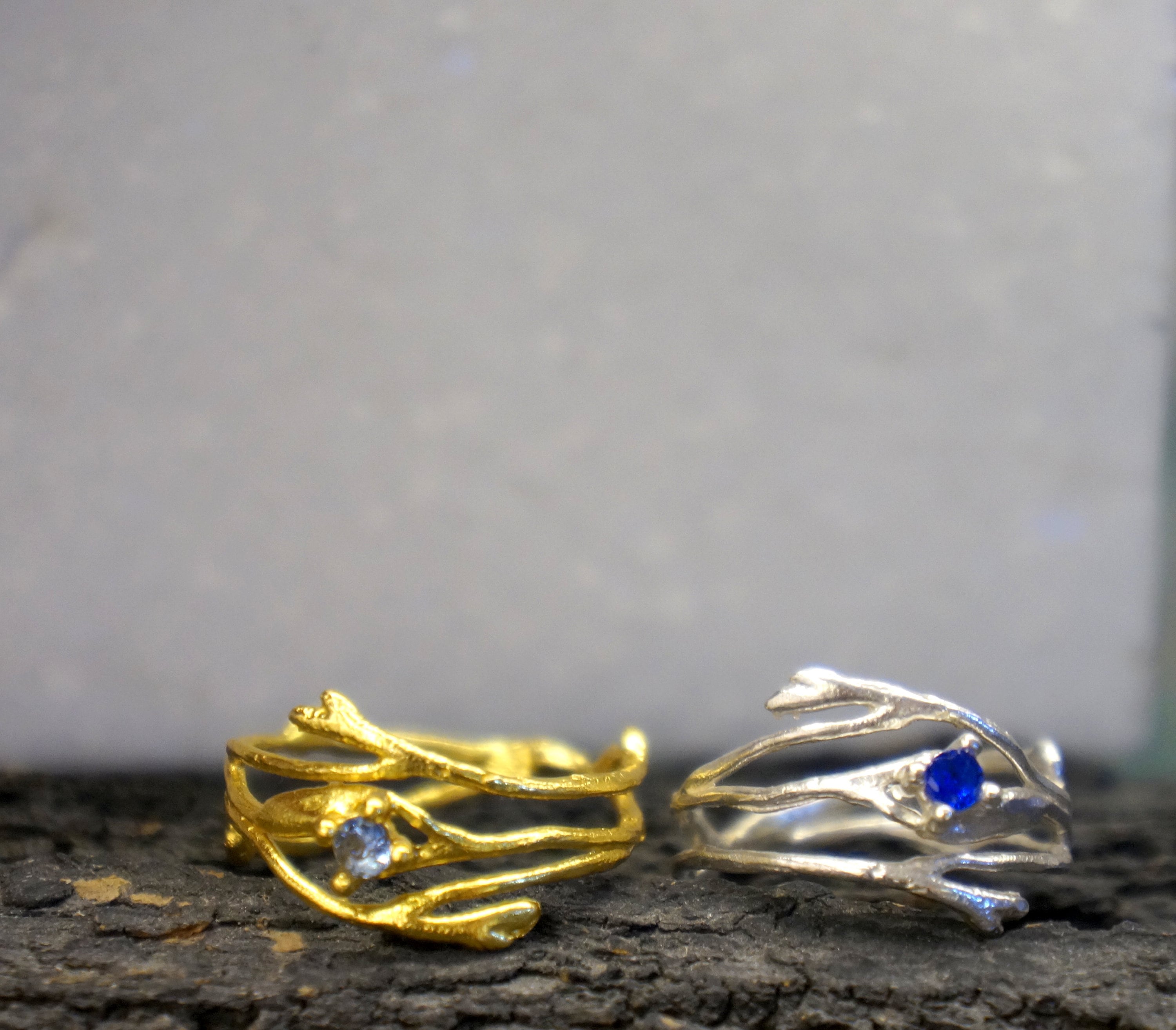 Two ornate rings with branches and gemstones are displayed against a gray background.