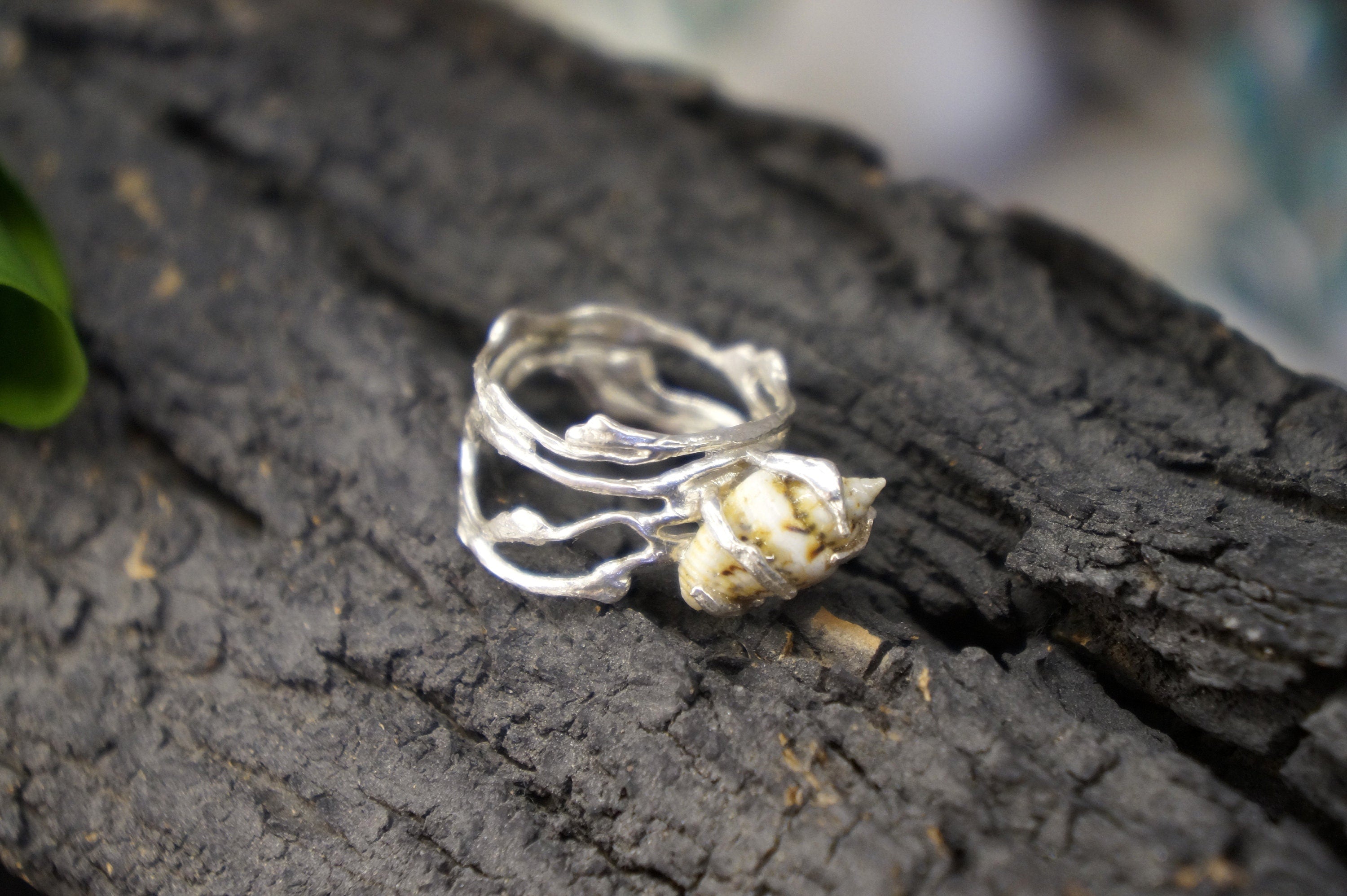 A silver ring with a shell-like design rests on a dark, textured surface.