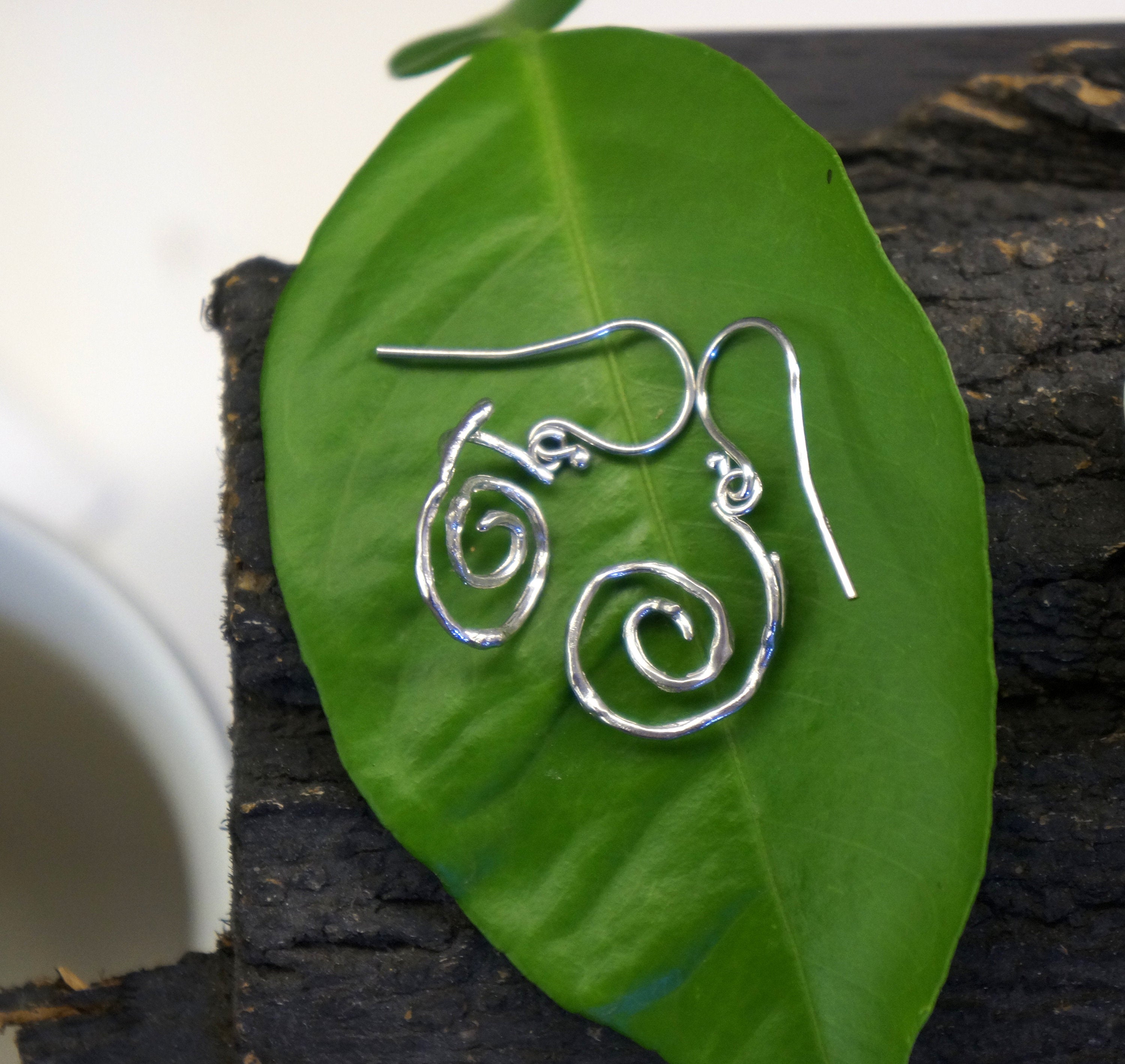 A pair of silver earrings with spiral designs rests on a green leaf.