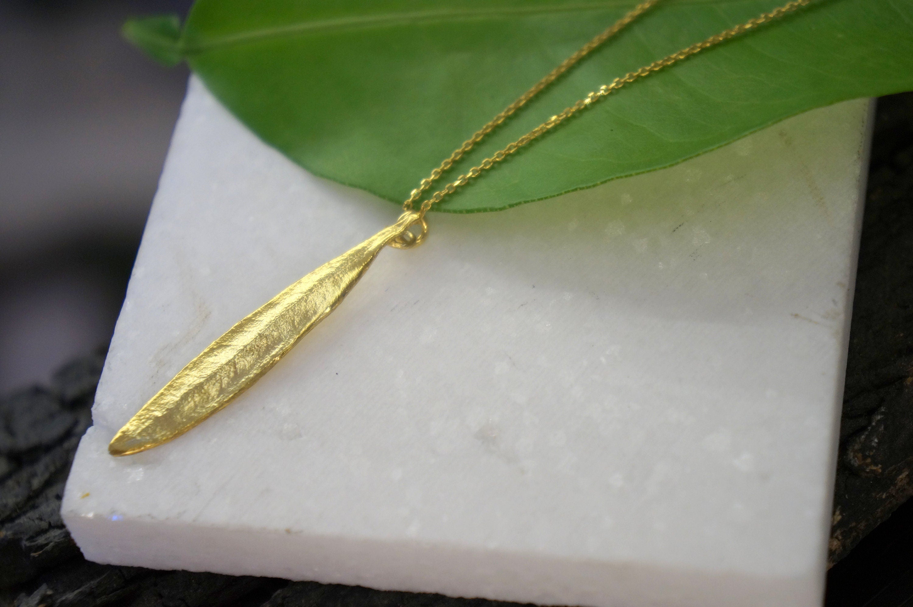 A gold necklace with a leaf pendant is displayed on a white marble surface, with a green leaf partially covering the necklace.