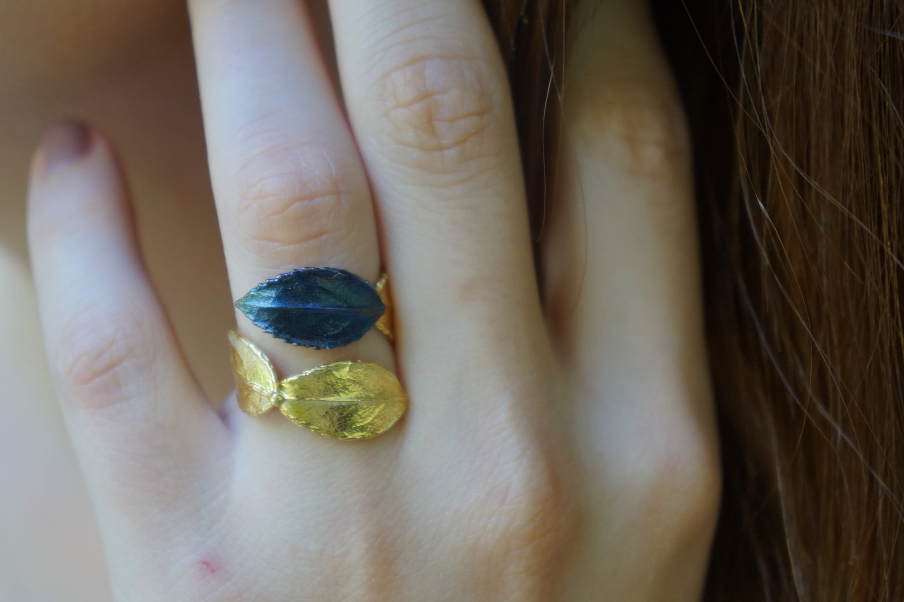 A close-up of a person's hand wearing a ring with two leaf-shaped gemstones, one blue and one gold.