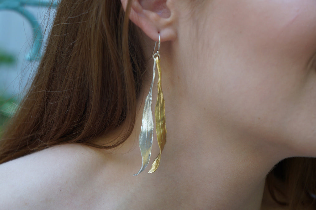 A close-up view of a person's ear wearing a pair of gold earrings with leaf-like designs.