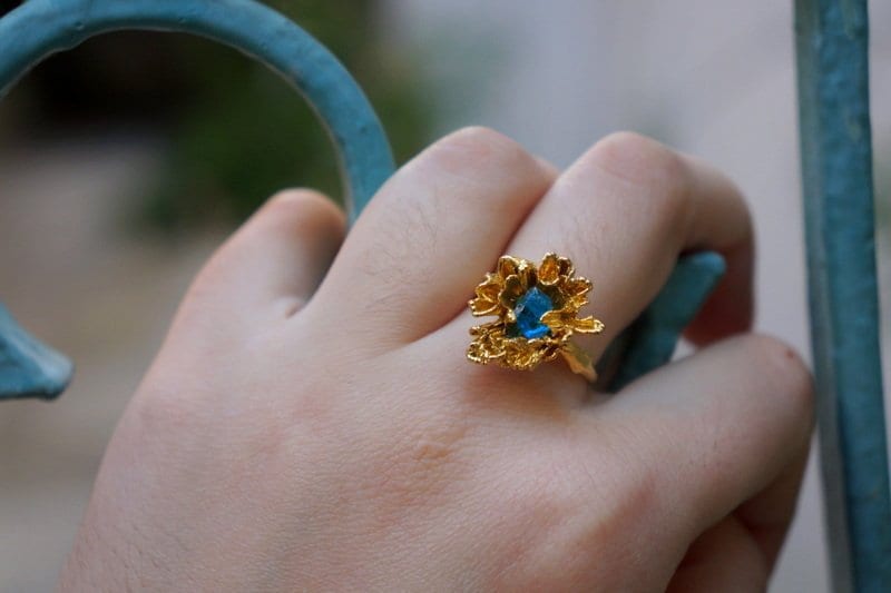 A close-up of a hand wearing a gold ring with a blue gemstone, holding a blue metal gate.