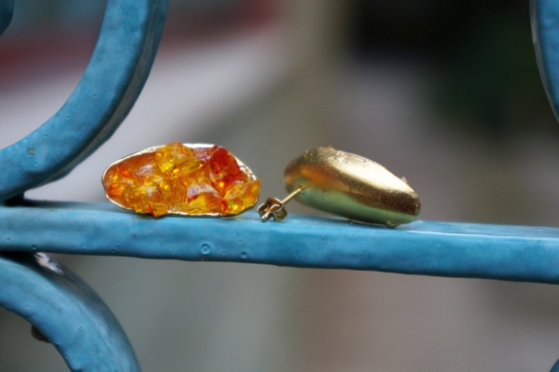 A pair of gold earrings with a vibrant orange stone is displayed on a blue metal railing.