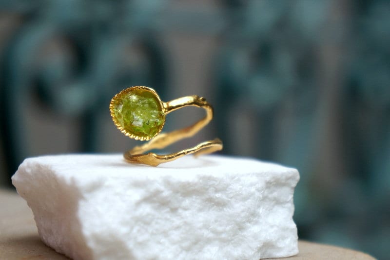 A gold ring with a green gemstone is displayed on a white marble surface against a blurred green background.
