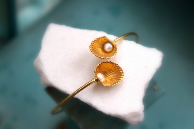 A gold bracelet with two shell-shaped charms, one with a pearl, resting on a white cloth.