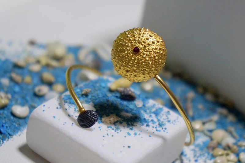 A gold ring with a round, textured ball-like design sits on a white surface, surrounded by blue and white pebbles.