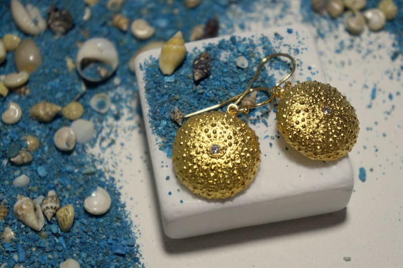 a pair of gold earrings with a textured design, placed on a white surface surrounded by various seashells and sand.