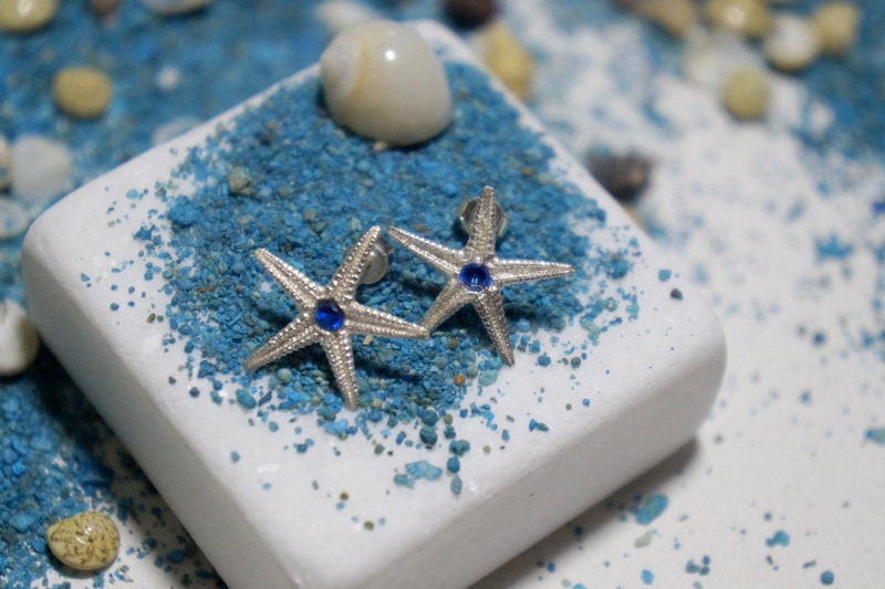 A pair of silver starfish earrings with blue gemstones, resting on a bed of blue sand or pebbles, creating a serene and nautical scene.