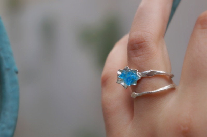 A close-up of a person's hand wearing a silver ring with a blue gemstone, with a blurred background.