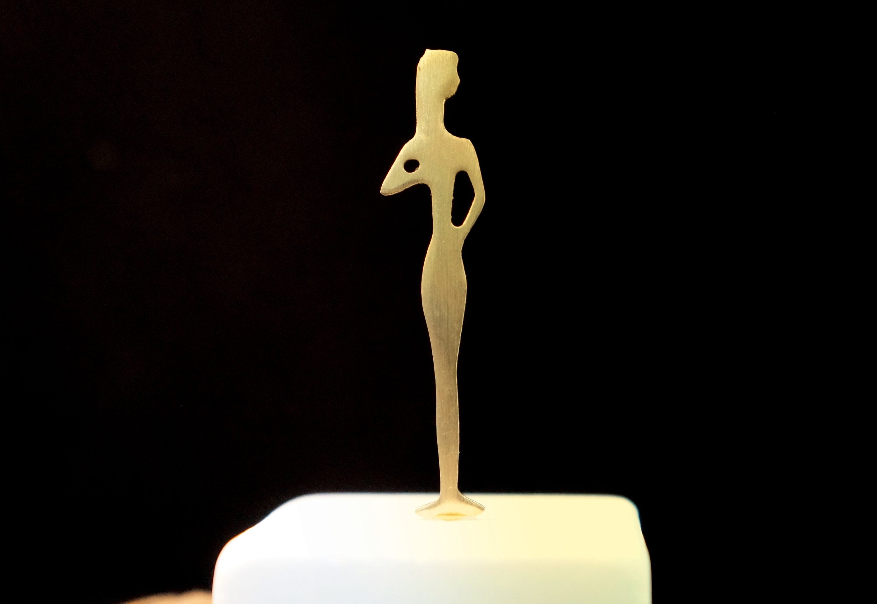 A golden statue of a woman is displayed on a white pedestal against a black background.
