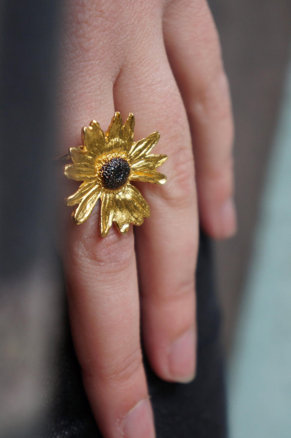 A close-up of a person's hand wearing a gold ring with a large, intricately designed flower as the centerpiece.