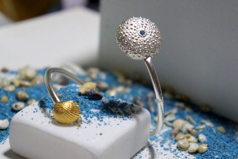 a silver ring with a round, textured ball at the top, placed on a white surface with blue sand or glitter.